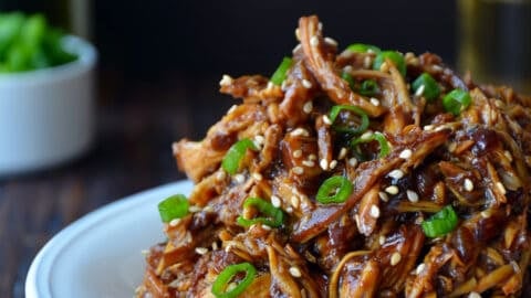 Shredded honey garlic chicken over brown rice in a bowl and garnished with sliced scallions and sesame seeds.