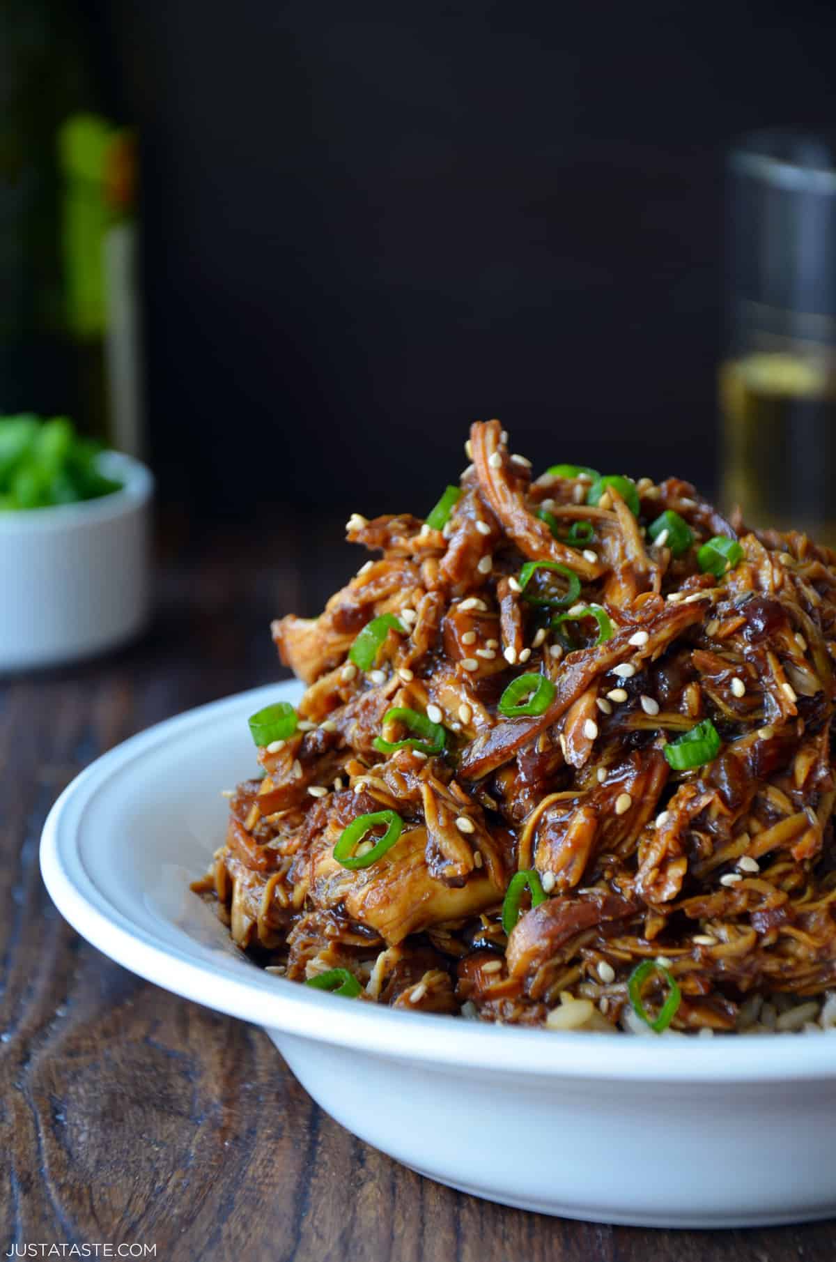 Shredded honey garlic chicken over brown rice in a bowl and garnished with sliced scallions and sesame seeds.