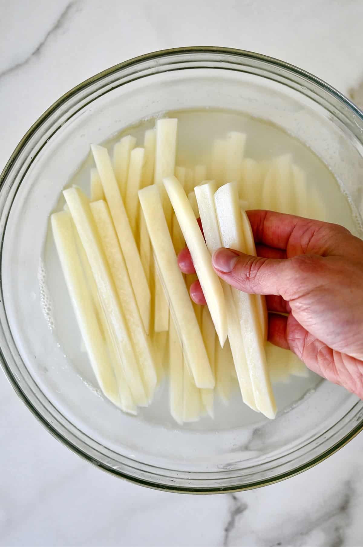 A hand holds slices of potatoes over a bowl of uncooked fries soaking in a water and white vinegar bath. 