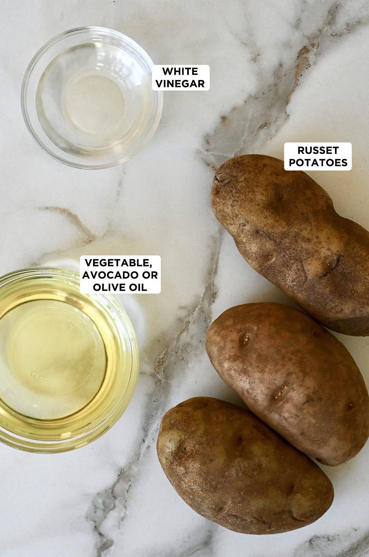 A small bowl filled with white vinegar next to a medium bowl with vegetable oil and three Russet potatoes.
