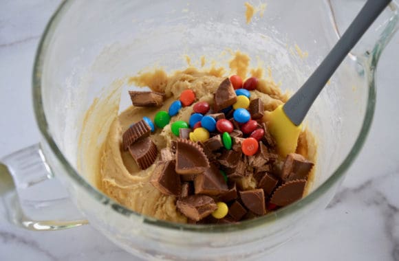 Spatula in glass bowl containing chopped Reese's Peanut Butter Cups, chopped Kit Kat Bars, and M&M's 