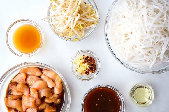 Various sizes of bowls containing bean sprouts, rice noodles, spices, soy sauce, diced chicken and chicken stock