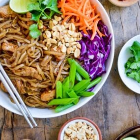 Easy Pad Thai with Chicken on a plate with chopsticks, cilantro, shredded carrots, shredded purple cabbage, scallions and peanuts