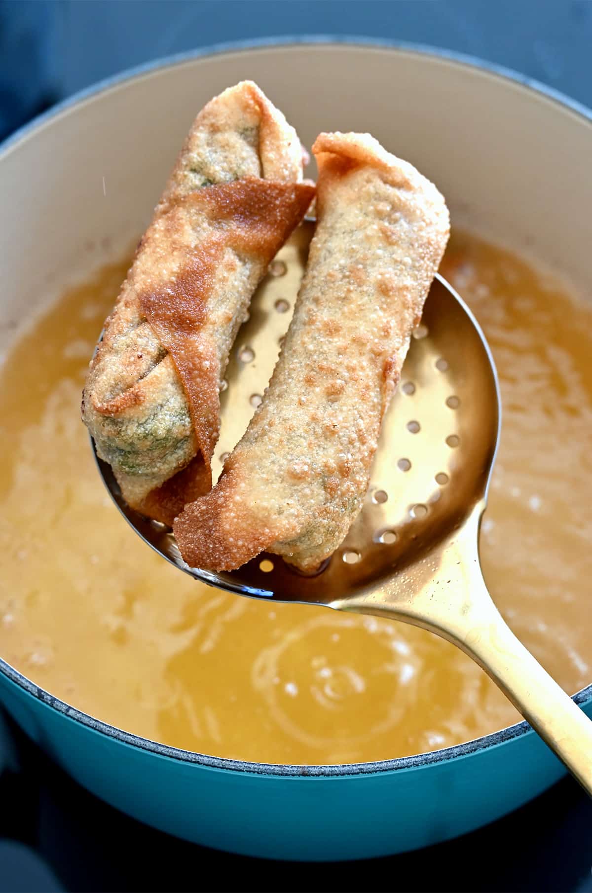 Two fried egg rolls on a slotted spoon over a large pot containing hot oil.