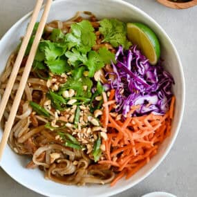 Chicken Pad Thai with chopped peanuts, sliced scallions, carrots and purple cabbage, garnished with a lime wedge and cilantro in a white bowl with chopsticks.
