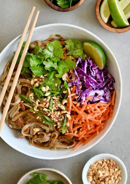 Chicken Pad Thai with chopped peanuts, sliced scallions, carrots and purple cabbage, garnished with a lime wedge and cilantro in a white bowl with chopsticks.
