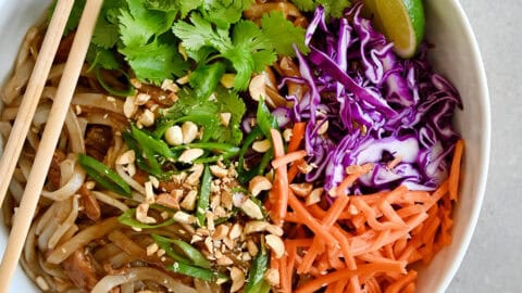 Chicken Pad Thai with chopped peanuts, sliced scallions, carrots and purple cabbage, garnished with a lime wedge and cilantro in a white bowl with chopsticks.