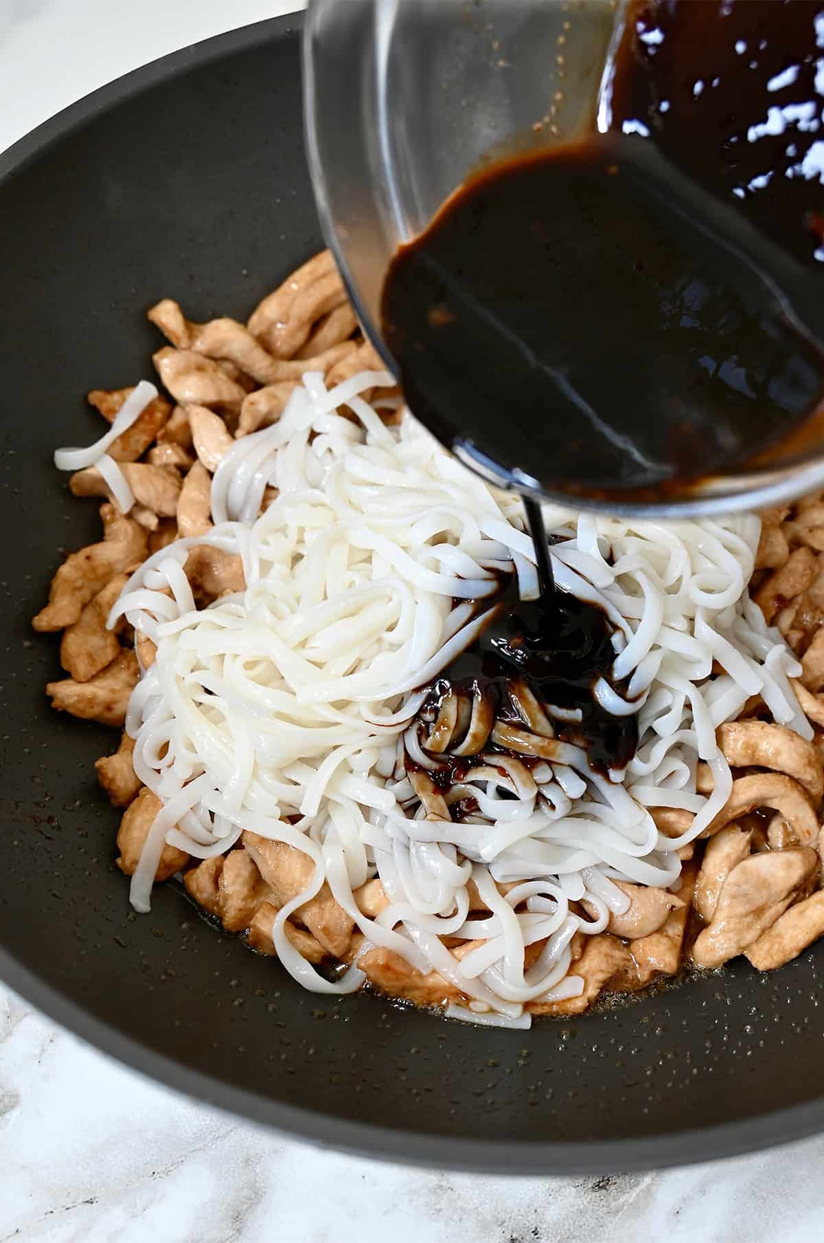Pad Thai sauce being poured from a small glass bowl over stir-fried chicken and rice noodles in a wok.
