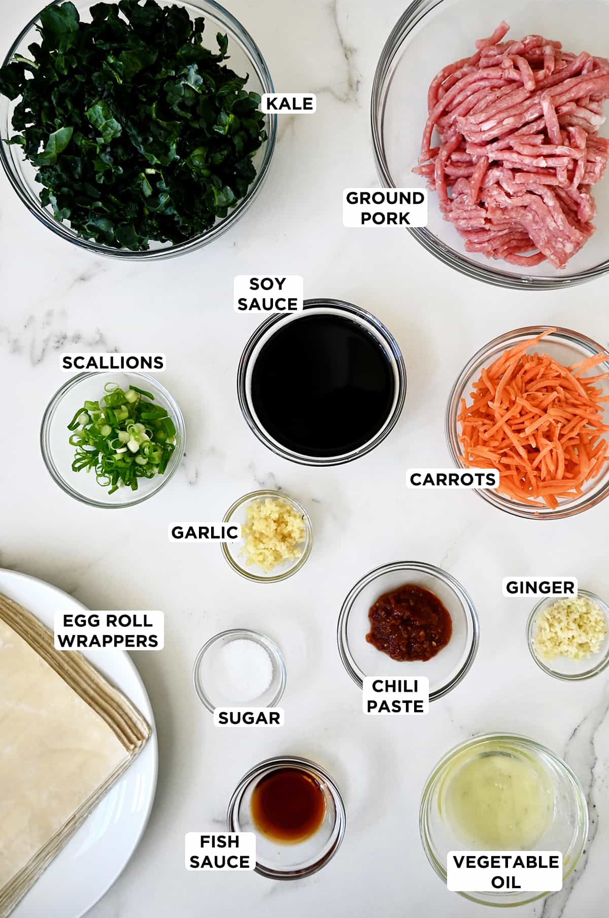 Various sizes of glass bowls containing pork egg roll and ginger soy dipping sauce ingredients, including ground pork, kale, soy sauce, shredded carrots, minced ginger, chili paste, sugar, minced garlic, scallions, fish sauce, vegetable oil and egg roll wrappers.