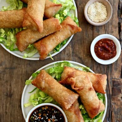 Two plates containing crispy pork egg rolls atop shredded lettuce and a small bowl filled with ginger soy dipping sauce.