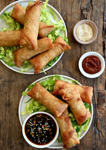 Two plates containing crispy pork egg rolls atop shredded lettuce and a small bowl filled with ginger soy dipping sauce.