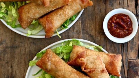 Two plates containing crispy pork egg rolls atop shredded lettuce and a small bowl filled with ginger soy dipping sauce.