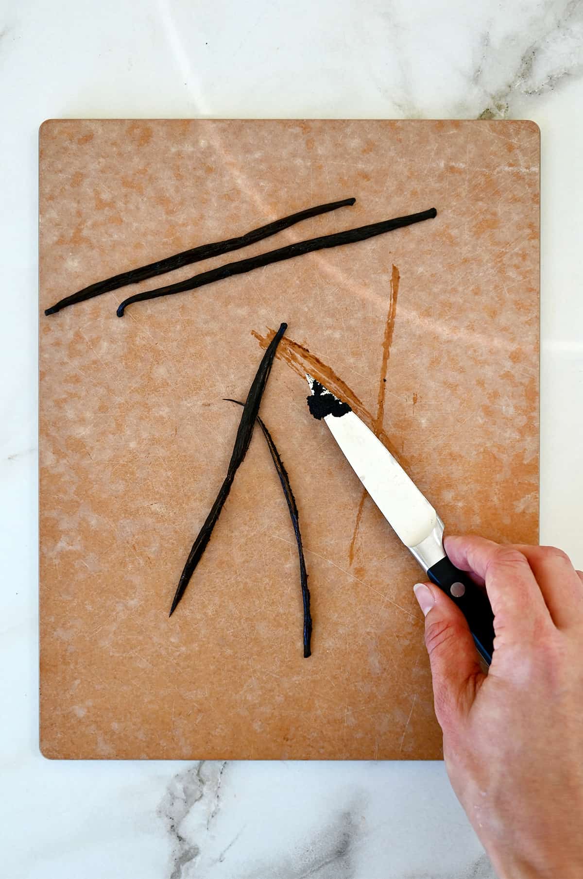 A hand holding a sharp knife scrapes vanilla beans out of a vanilla bean pod.