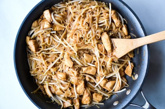 Wooden spoon in skillet with bean sprouts, rice noodles and diced chicken