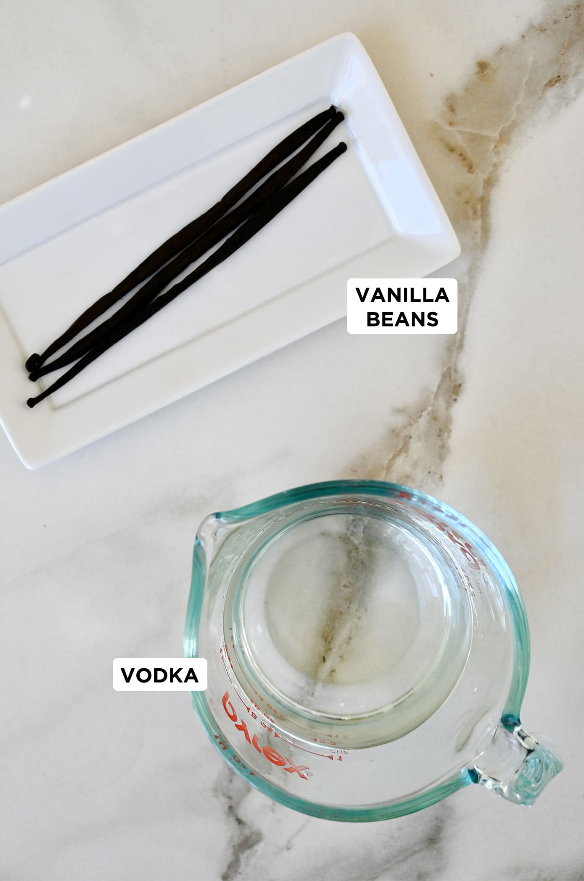 Vanilla beans on a white plate next to a liquid measuring cup containing vodka.