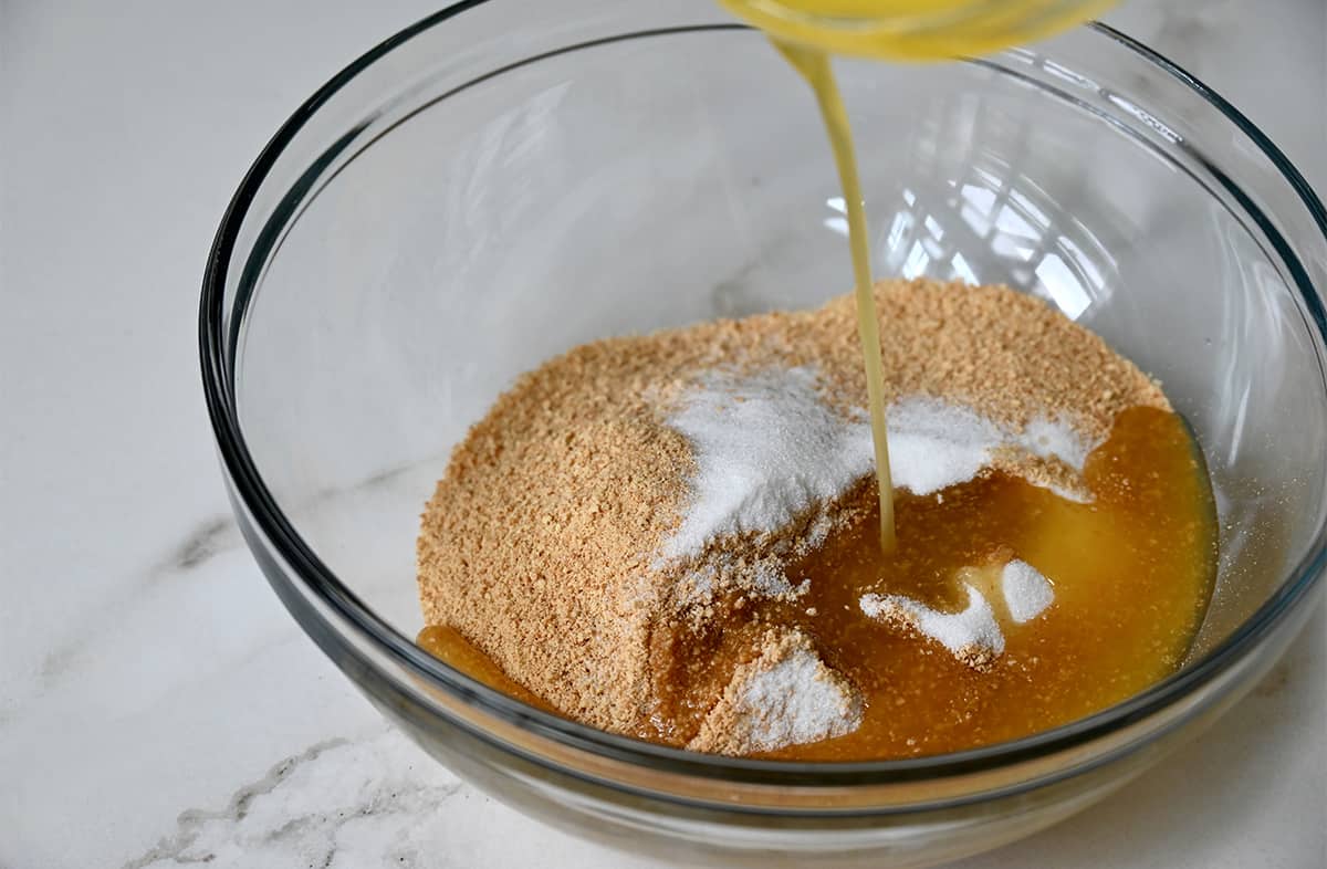 Melted butter being poured atop crushed graham crackers and sugar in a clear bowl.
