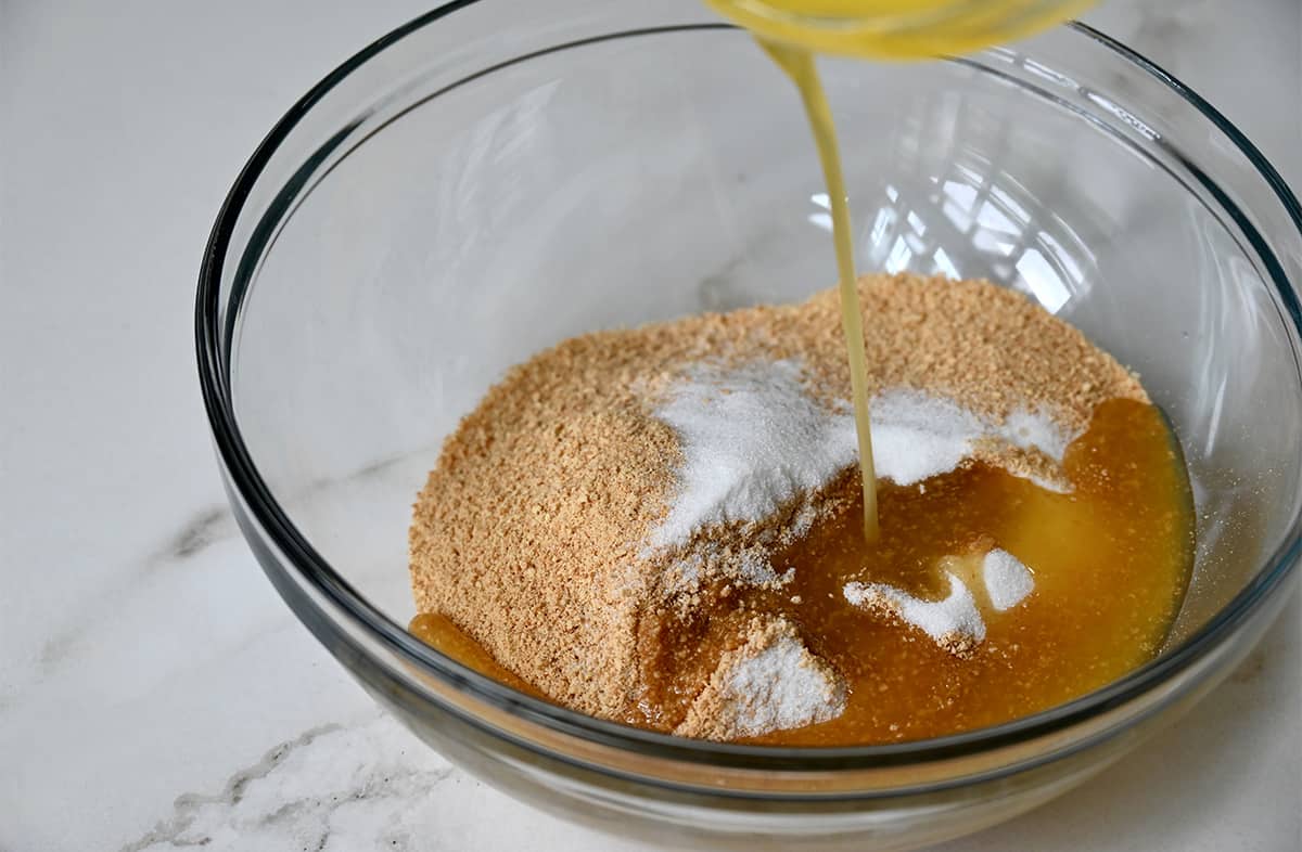 Easy No-Bake Cheesecake from JustaTaste.com #recipe Melted butter being poured atop crushed graham crackers and sugar in a clear bowl.