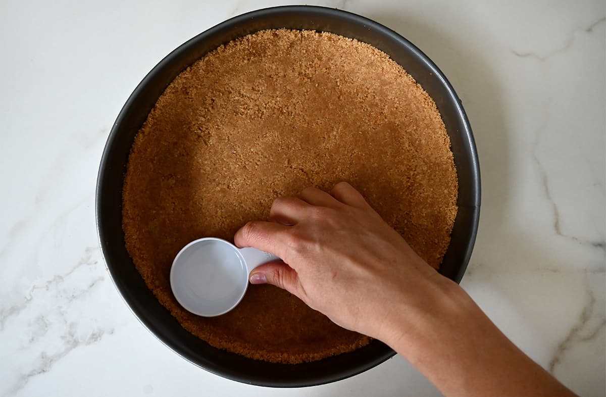 A top-down view of a hand holding a measuring cup pressing graham cracker crumbs into a springform pan.