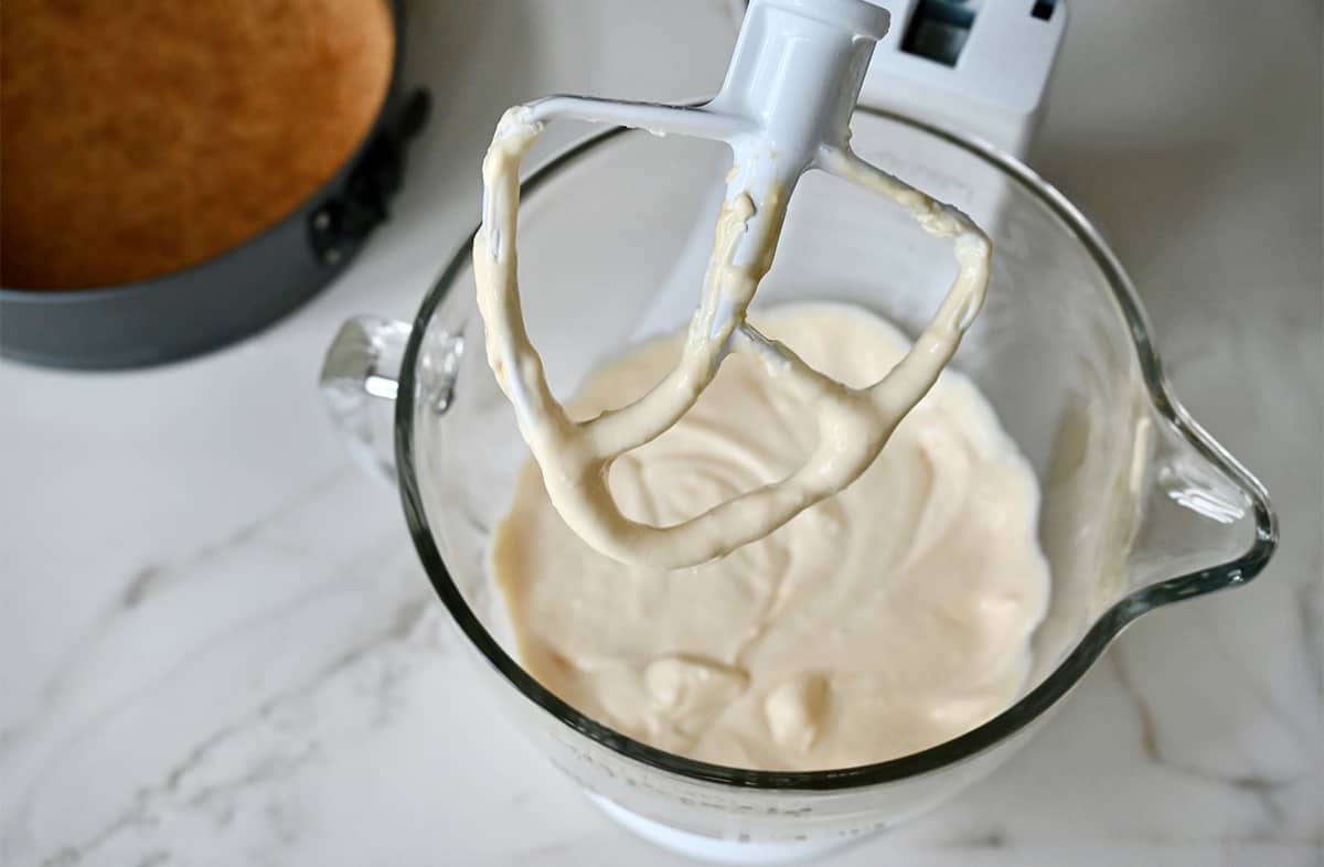 The paddle attachment of a stand mixer over a clear stand mixer bowl containing creamy no-bake cheesecake filling.