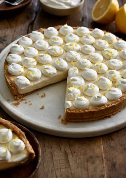 A no-bake cheesecake with a graham cracker crust topped with dollops of whipped cream and lemon zest with one slice missing.
