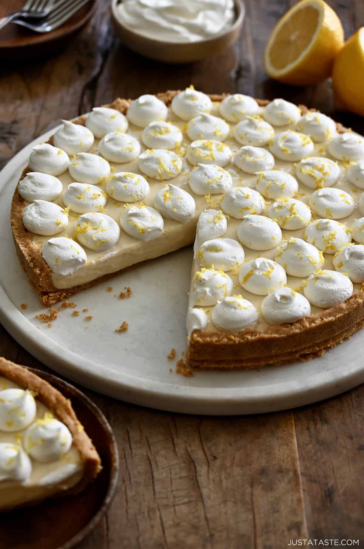 A no-bake cheesecake with a graham cracker crust topped with dollops of whipped cream and lemon zest with one slice missing.