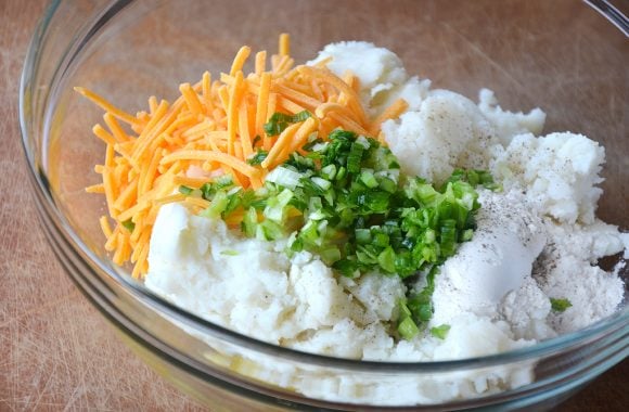 A glass bowl containing leftover mashed potatoes, cheddar cheese and scallions