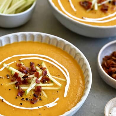 Two bowls containing butternut squash apple soup garnished with apple matchsticks, crispy pancetta and a drizzle of sour cream.