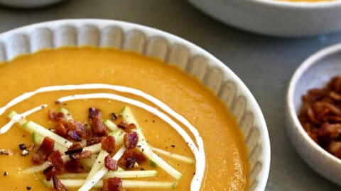 Two bowls containing butternut squash apple soup garnished with apple matchsticks, crispy pancetta and a drizzle of sour cream.