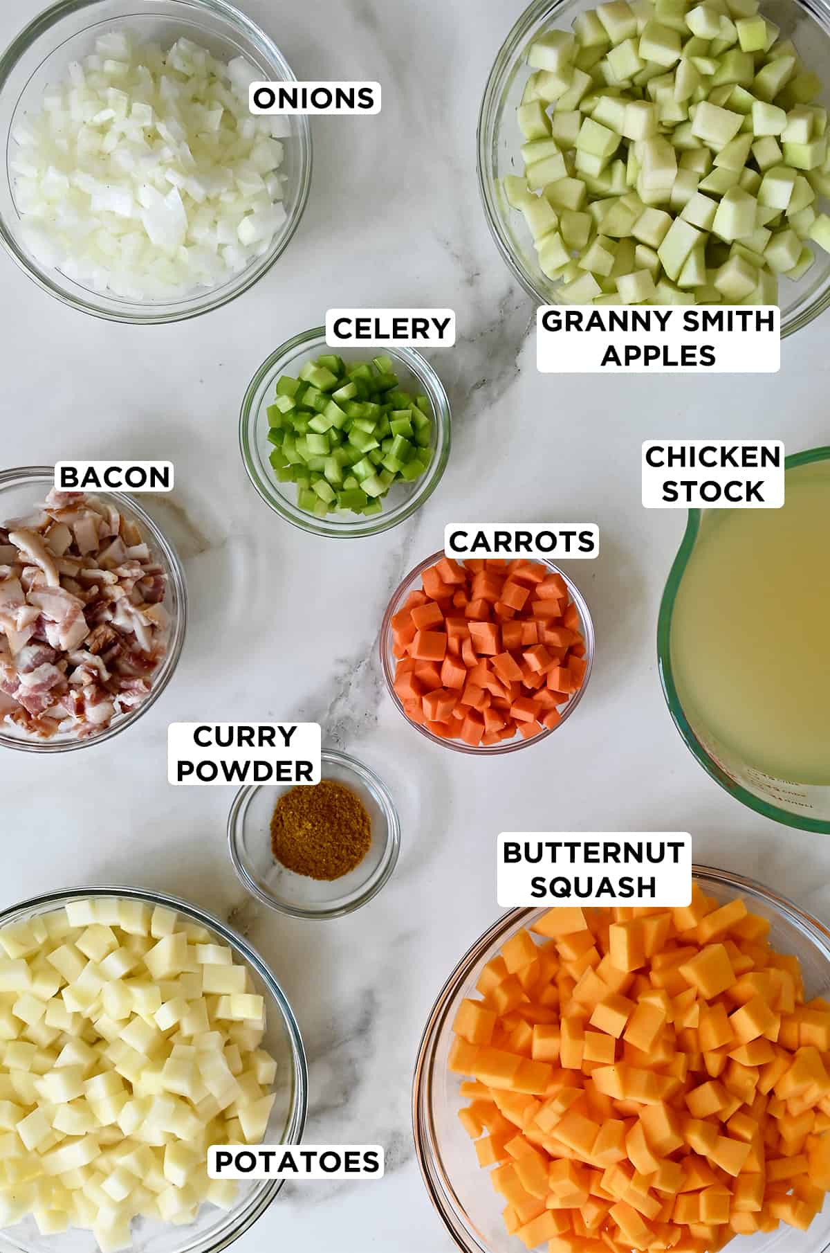 Various sizes of clear bowls containing chopped onion, chopped Granny Smith apples, chicken stock, cubed butternut squash, chopped carrots, curry powder, chopped celery, bacon and chopped Yukon gold potatoes.