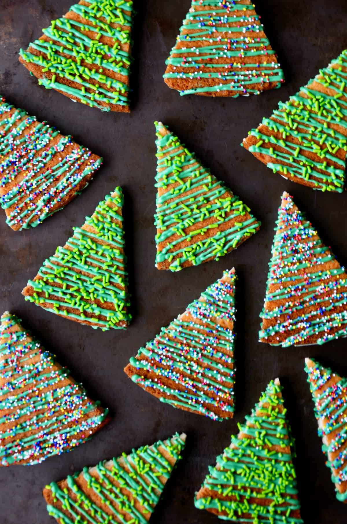 Christmas tree shortbread cookies decorated with green icing and sprinkles are scattered on a dark-colored countertop.