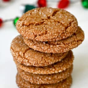 A stack of sugar-dusted ginger cookies are on a white surface, with red and green decorations in the background.