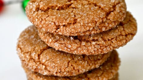 A stack of sugar-dusted ginger cookies are on a white surface, with red and green decorations in the background.