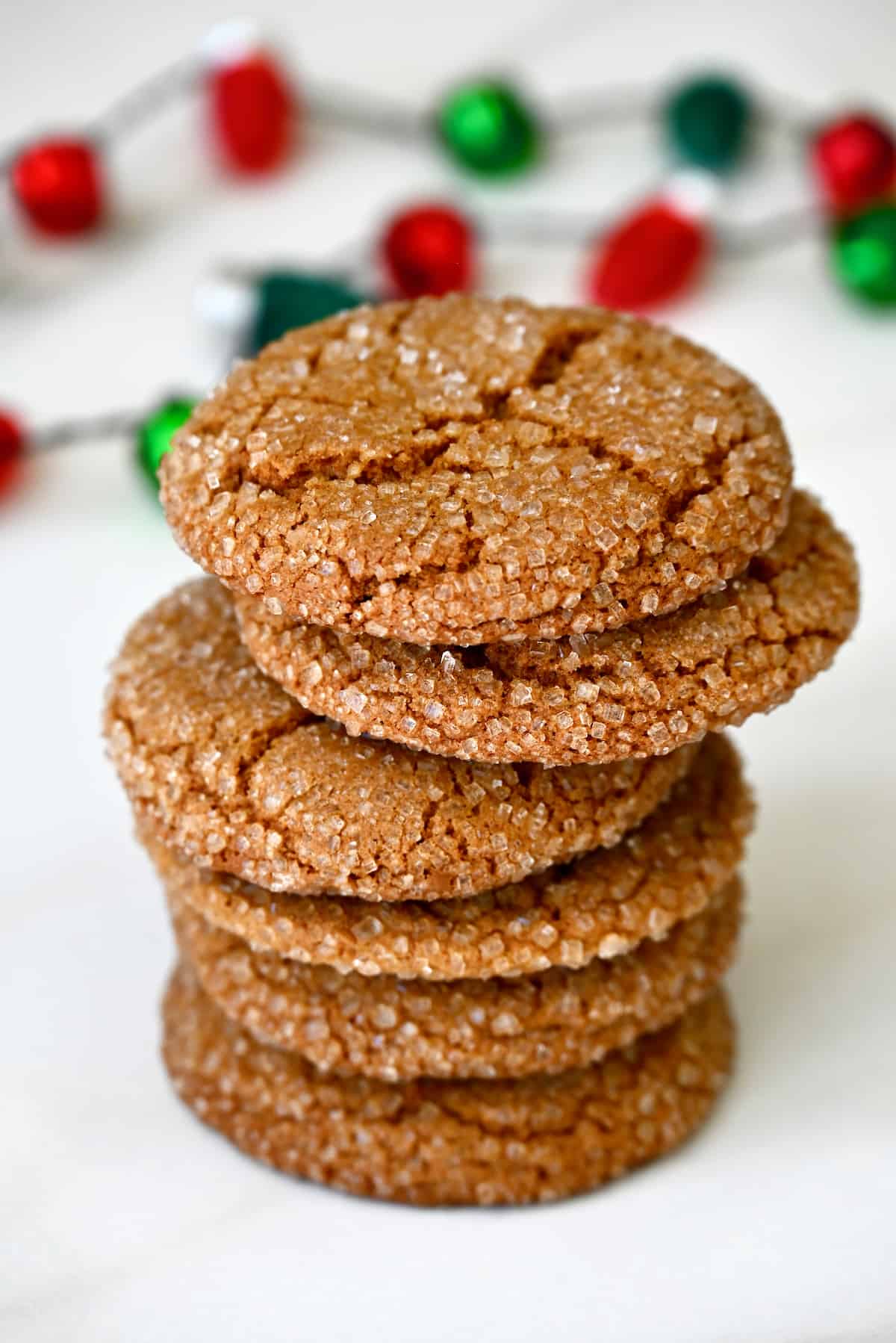 A stack of sugar-dusted ginger cookies are on a white surface, with red and green decorations in the background.