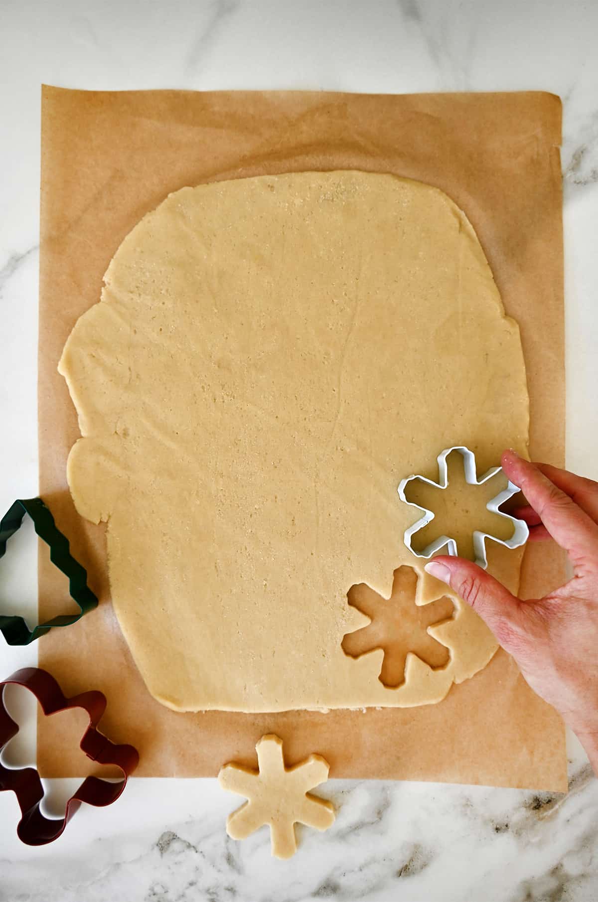 Rolled out dough on a sheet of parchment paper being cut into snowflake shapes with a gingerbread man and Christmas tree cookie cutter next to it.