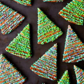 Christmas tree shortbread cookies decorated with green icing and sprinkles are scattered on a dark-colored countertop.