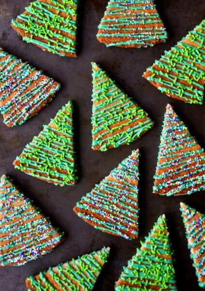 Christmas tree shortbread cookies decorated with green icing and sprinkles are scattered on a dark-colored countertop.