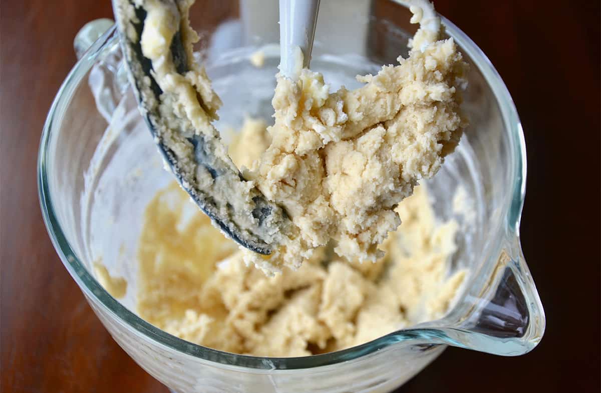 Shortbread cookie dough is in a glass mixing bowl with the dough-covered paddle of a stand mixer hanging over it.