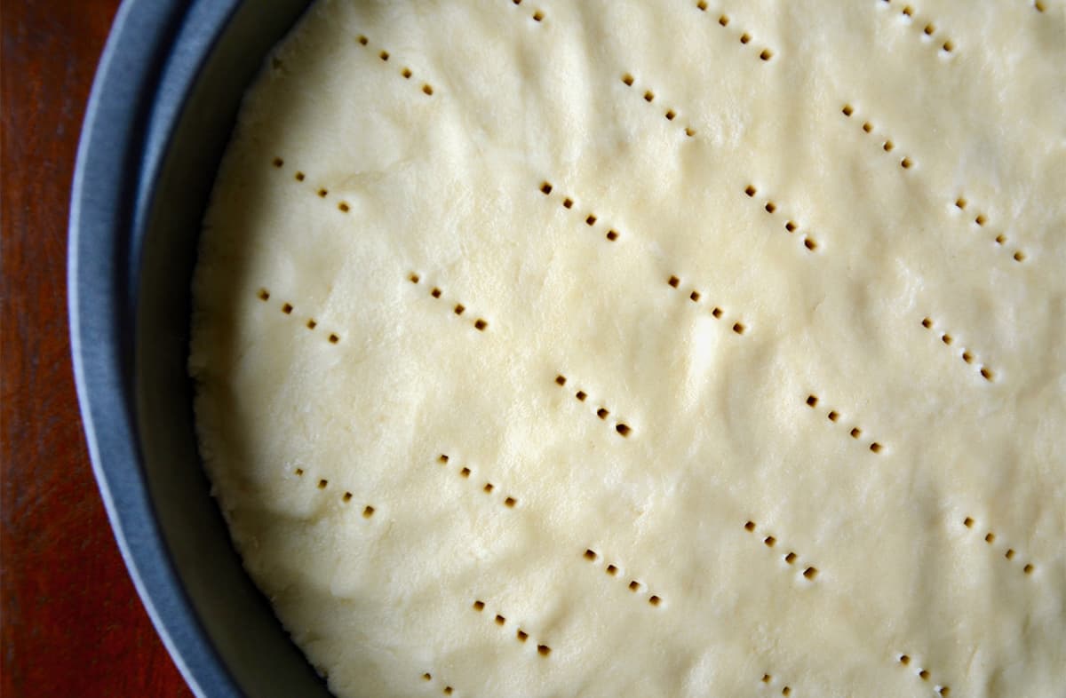 Shortbread cookie dough is pressed into a round cake pan with neat rows of prick marks from a fork.