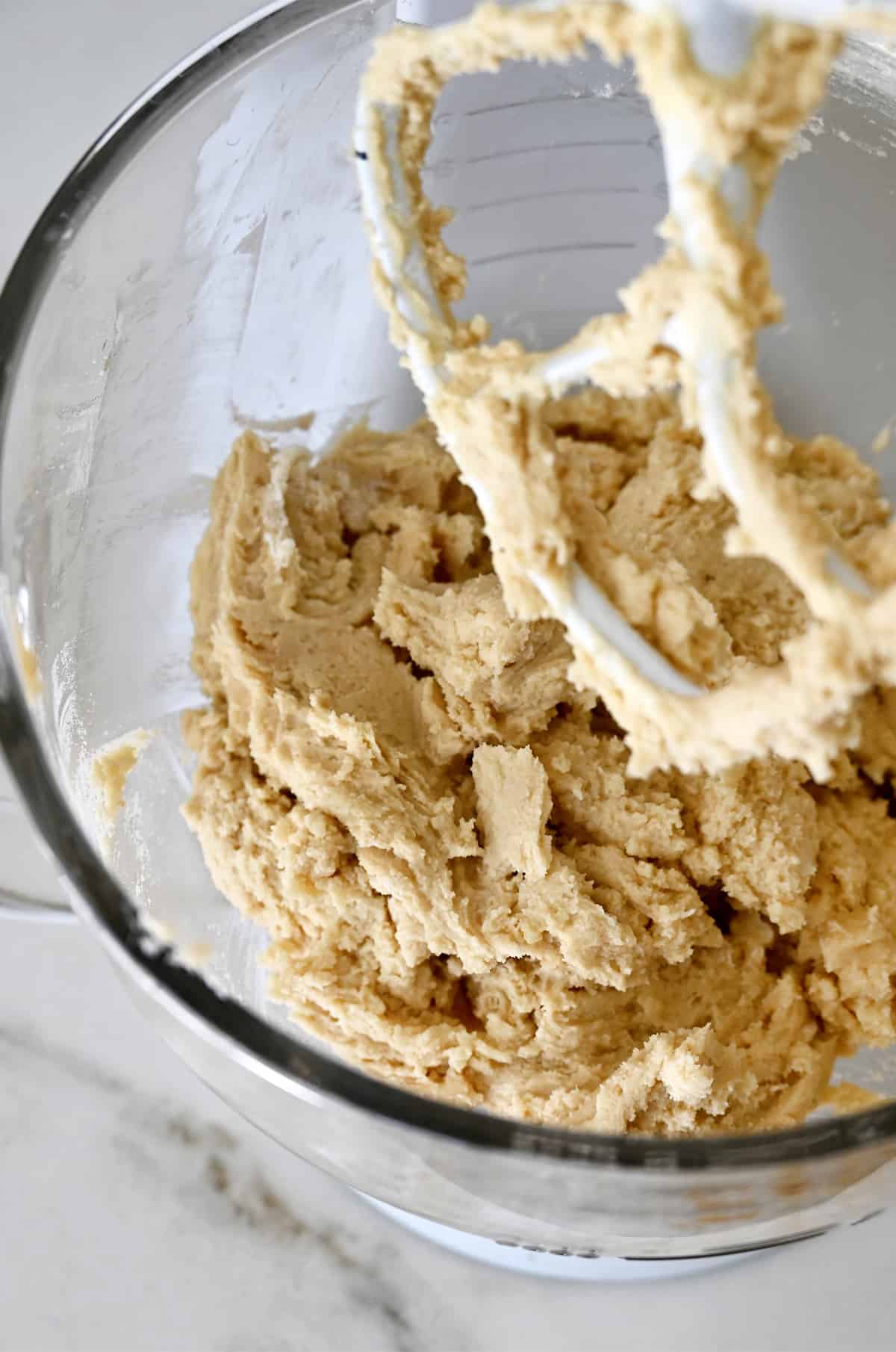 Sugar cookie dough covering the paddle attachment of a stand mixer above the glass bowl of the mixer containing more cookie dough.