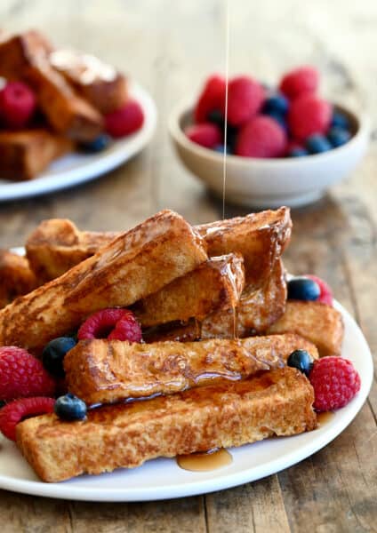 Several cinnamon French toast sticks on a white plate with fresh raspberries and blueberries all being drizzled with warm maple syrup.