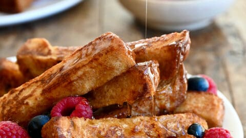 Several cinnamon French toast sticks on a white plate with fresh raspberries and blueberries all being drizzled with warm maple syrup.