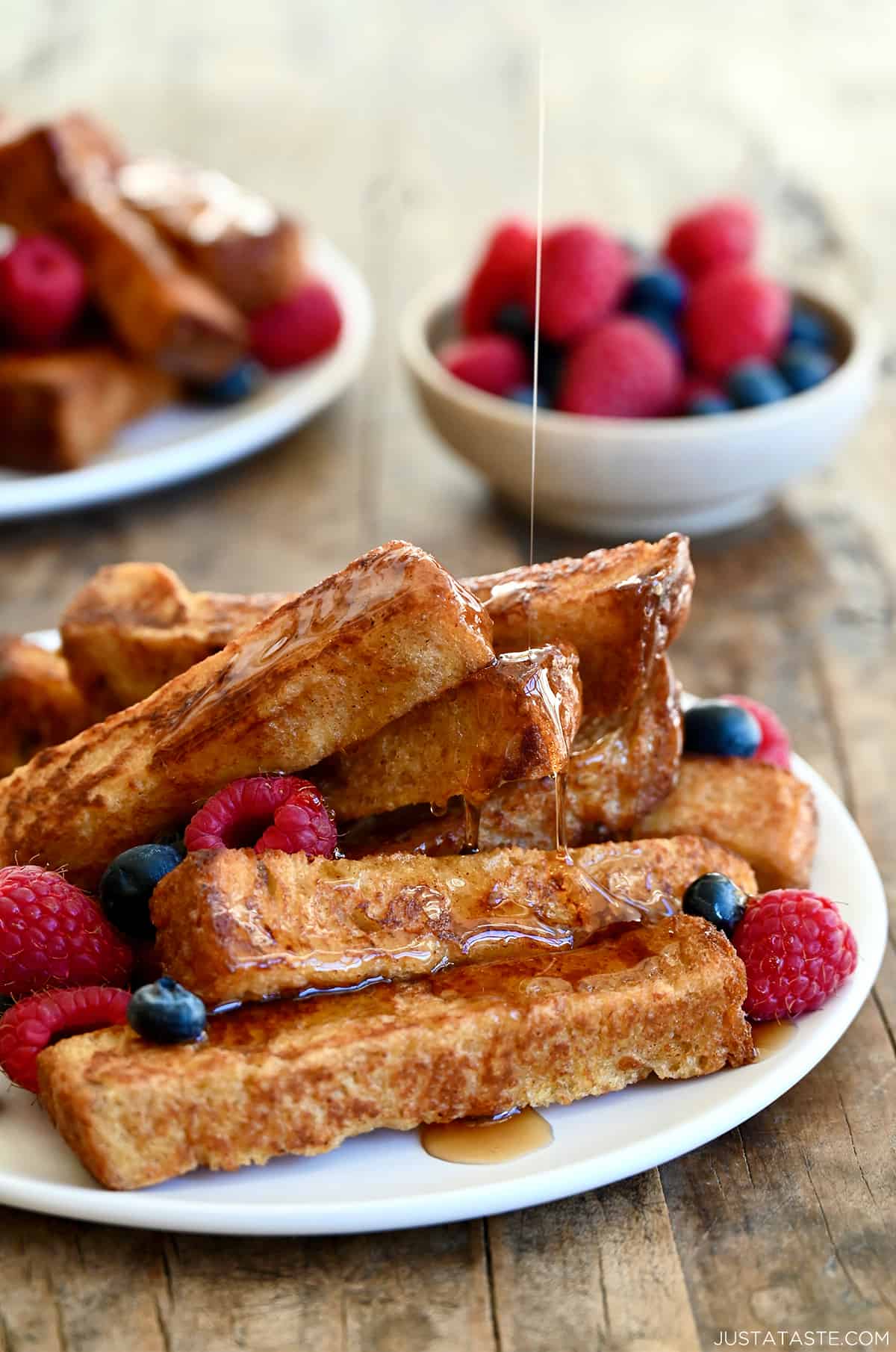 Several cinnamon French toast sticks on a white plate with fresh raspberries and blueberries all being drizzled with warm maple syrup. 
