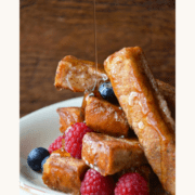 A bowl containing a stack of Cinnamon French Toast Sticks drizzled with maple syrup and garnished with fresh raspberries and blueberries.