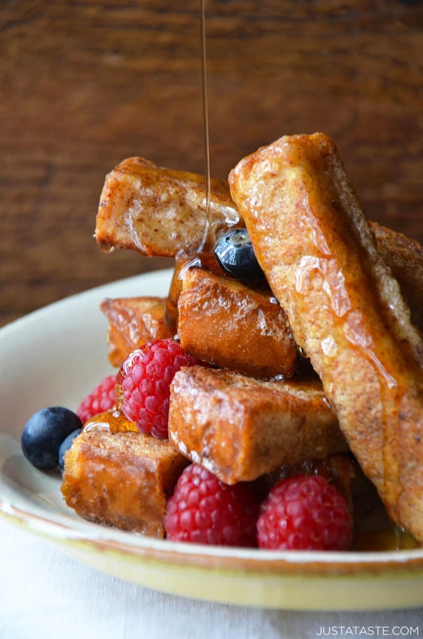 White plate containing a stack of Easy Cinnamon French Toast Sticks topped with fresh raspberries and blueberries, and maple syrup