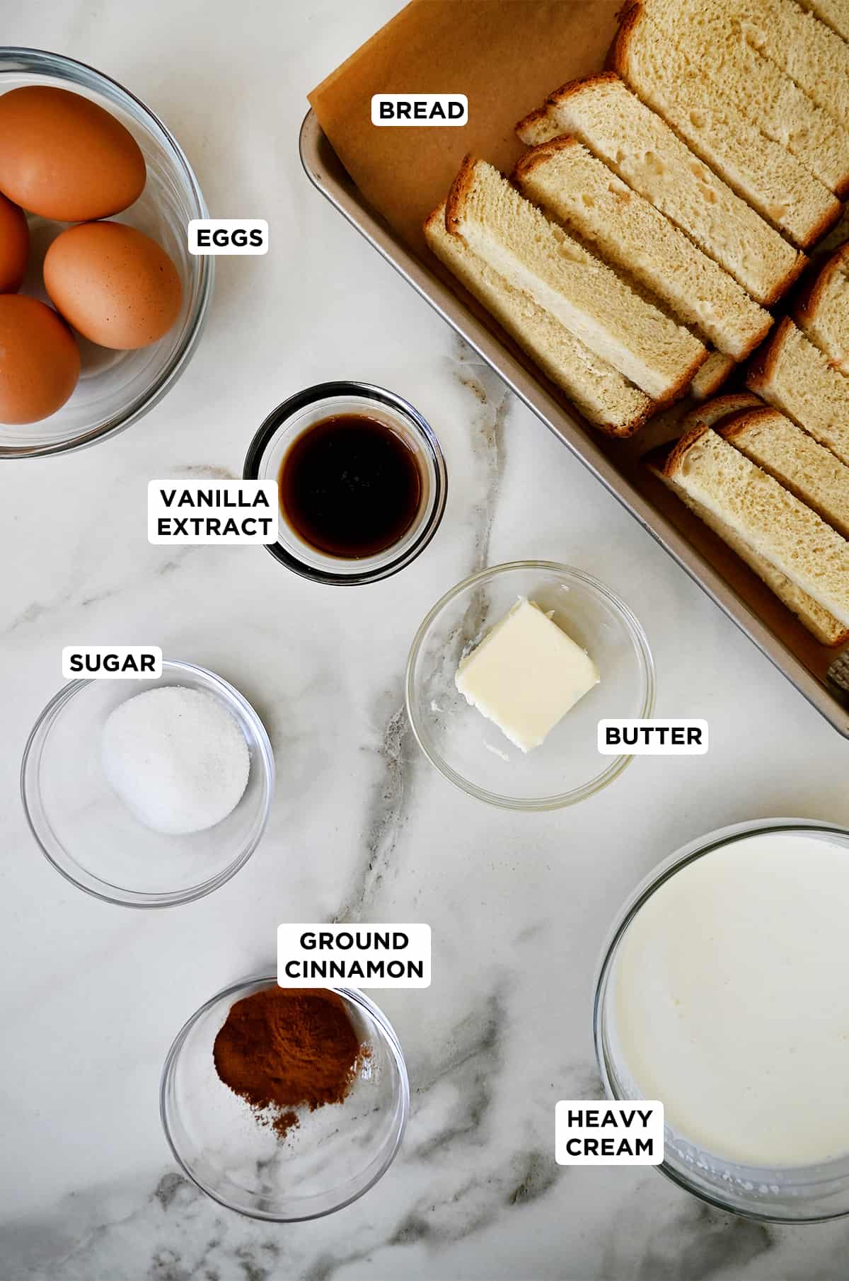 Batons of Texas toast on a cutting board next to various sizes of glass bowls containing three eggs, vanilla extract, butter, sugar, ground cinnamon and heavy cream.