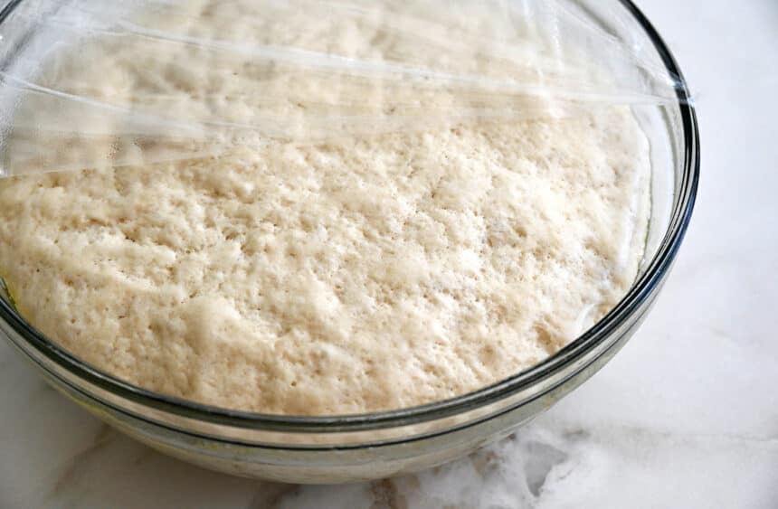 Proofed dough in a clear bowl covered with plastic wrap