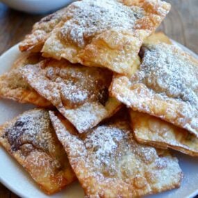 A white plate containing a pile of chocolate wontons covered with powdered sugar