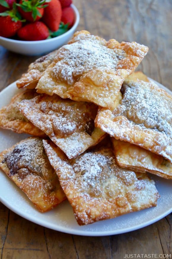 A white plate containing a pile of chocolate wontons covered with powdered sugar