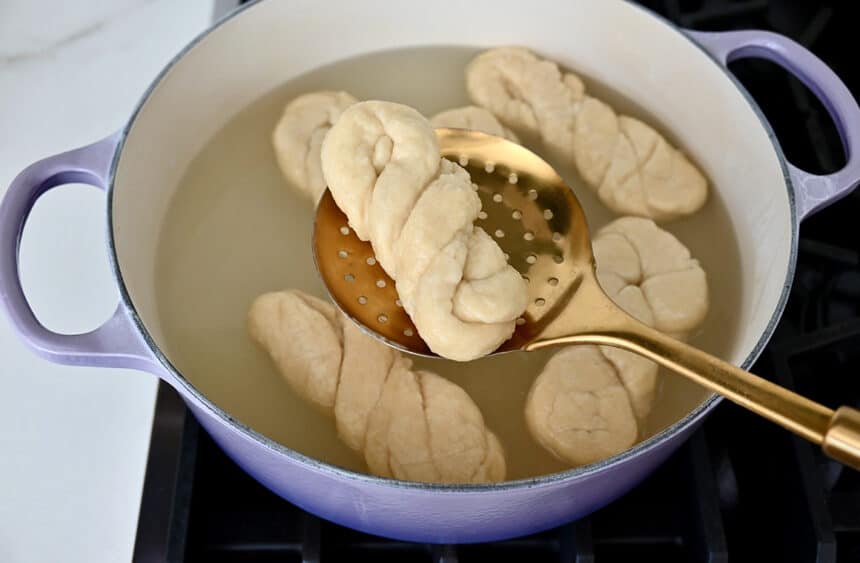 A slotted spoon holds a pretzel twist over a stockpot containing a baking soda-pretzel bath