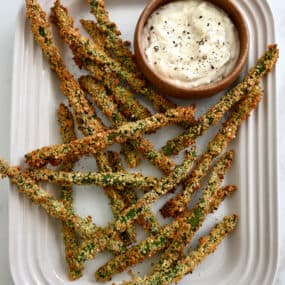 A top-down view of Baked Asparagus Fries on a white serving platter next to a small bowl containing roasted garlic aioli